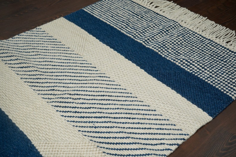 May include: A white and blue woven rug with a geometric pattern. The rug has a fringe border and is made of natural fibers.