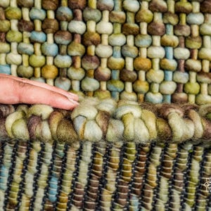 May include: Close-up of a woven rug with a textured surface. The rug is made of wool and has a variety of colors, including brown, green, and blue. The rug has a fringe border.