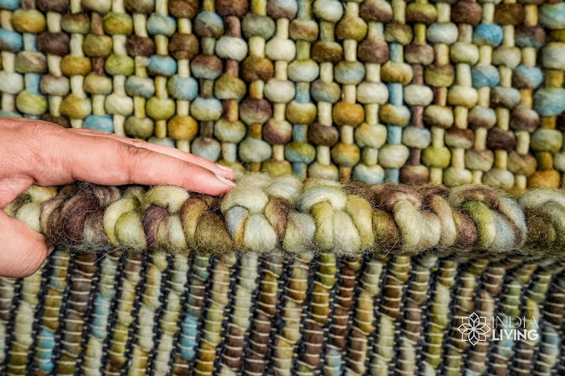 May include: Close-up of a woven rug with a textured surface. The rug is made of wool and has a variety of colors, including brown, green, and blue. The rug has a fringe border.