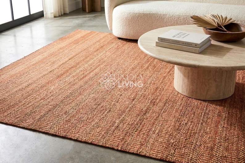 May include: A rectangular area rug featuring a woven herringbone pattern in shades of terracotta and natural tan. The rug has a textured, fibrous surface appearance and is laid flat on a smooth floor surface, extending under a round stone coffee table.