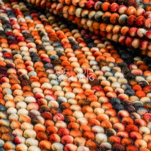 May include: Close-up of a colorful rug with a textured, bumpy surface. The rug features a pattern of small, round, wool-like elements in shades of orange, red, white, and blue. The rug is likely handmade and adds warmth and texture to a room.