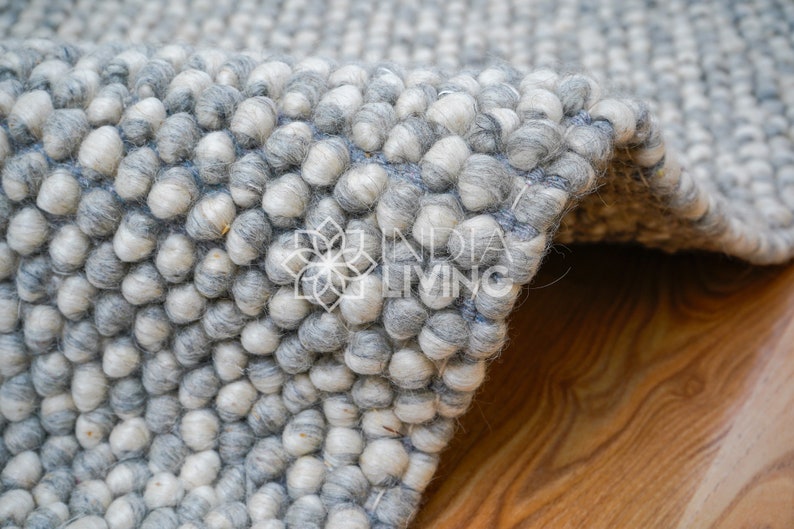 May include: Close-up of a gray and white fluffy rug made of small, round balls of wool. The rug is folded in half, showing the texture of the material.