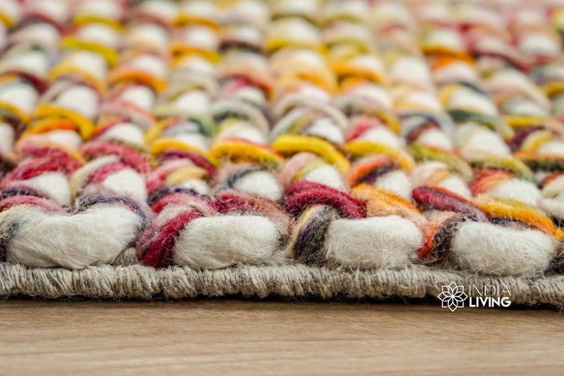 May include: A thick, hand-woven rug featuring a chunky, braided texture made from multi-colored yarn in shades of white, yellow, orange, red, and dark grey. The rug has a sturdy, stitched border edge and includes the India Living logo in the corner.