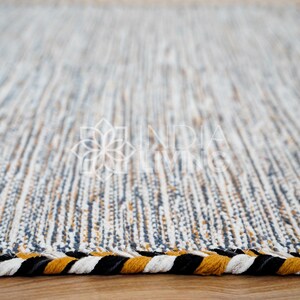 May include: A rectangular area rug featuring a woven texture with vertical stripes in shades of white, blue, and mustard yellow. The edges are finished with a thick, braided border consisting of alternating black, white, and mustard yellow strands.