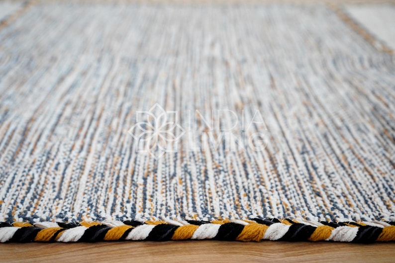 May include: A rectangular area rug featuring a woven texture with vertical stripes in shades of white, blue, and mustard yellow. The edges are finished with a thick, braided border consisting of alternating black, white, and mustard yellow strands.