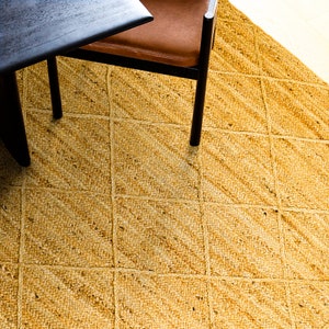 May include: A close-up of a woven jute rug with a diamond pattern in a light yellow color. The rug is made of natural fibers and has a rustic, earthy feel.