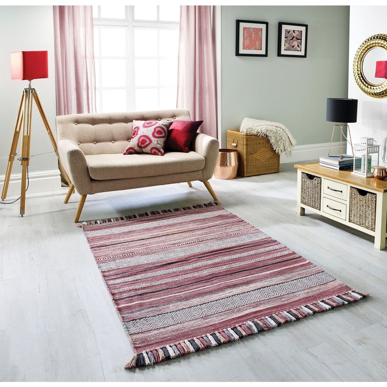 May include: A rectangular rug with horizontal stripes in shades of red, gray, and white, with a fringed edge. The rug is placed on a light-colored wooden floor in a living room setting. A beige sofa and decorative pillows are visible in the background.