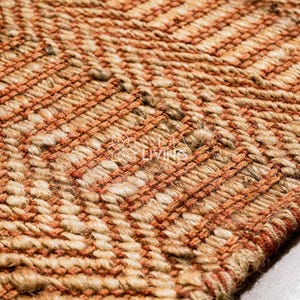 May include: A woven rug featuring a textured herringbone pattern in alternating bands of natural tan jute and burnt orange fibers. The surface displays a thick, braided construction with a visible logo reading "MY LIVING" in the center of the weave.