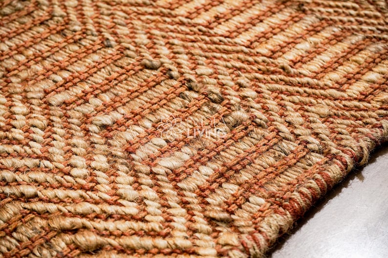 May include: A woven rug featuring a textured herringbone pattern in alternating bands of natural tan jute and burnt orange fibers. The surface displays a thick, braided construction with a visible logo reading "MY LIVING" in the center of the weave.