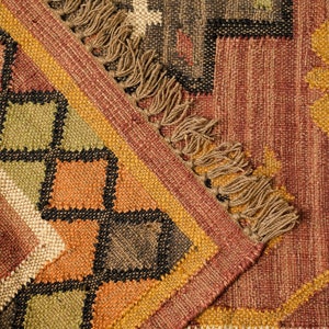 May include: A close-up of a woven rug with a geometric pattern in shades of brown, orange, and green. The rug has a fringe border.