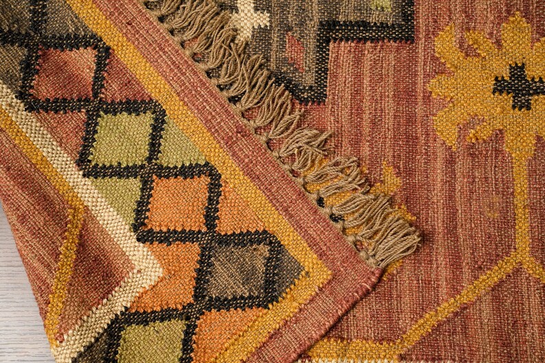 May include: A close-up of a woven rug with a geometric pattern in shades of brown, orange, and green. The rug has a fringe border.