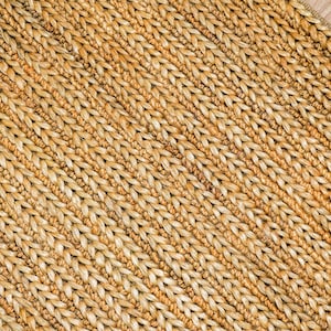 May include: Close-up of a woven rug with a natural, light brown color. The rug has a textured surface with a diagonal pattern. The weave is tight and appears durable, suitable for home decor.