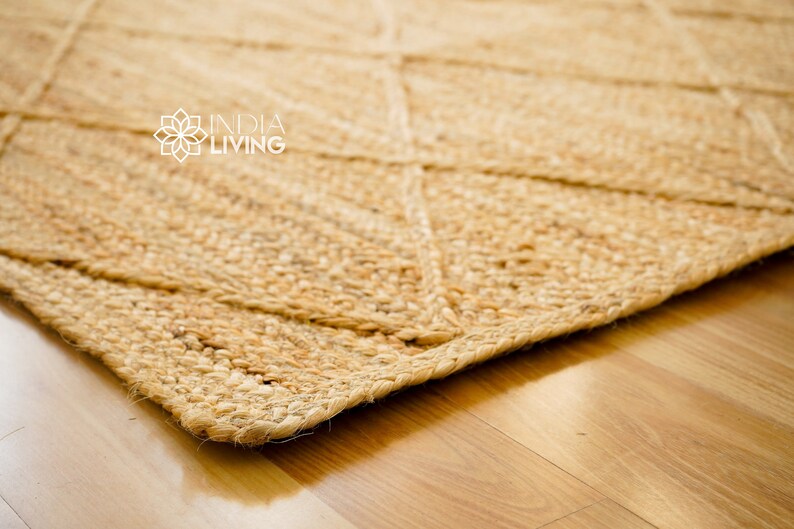 May include: A close-up of a natural fiber rug with a diamond pattern. The rug is a light brown color and has a rough texture. The rug is made of jute and is perfect for adding a natural touch to any room. INDIA LIVING is printed in white text on the rug.