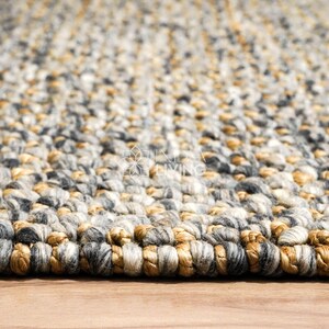 Puede incluir: Una alfombra gruesa tejida a mano con una textura trenzada y voluminosa. La alfombra est&aacute; compuesta por una mezcla de hilos en tonos gris carb&oacute;n, gris claro, crema y marr&oacute;n dorado c&aacute;lido, creando un aspecto variado y multicolor en toda la pieza.
