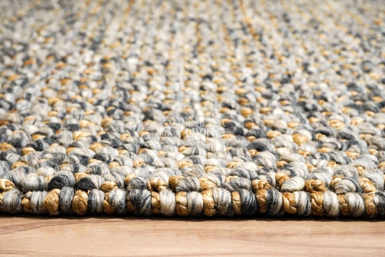 Puede incluir: Una alfombra gruesa tejida a mano con una textura trenzada y voluminosa. La alfombra est&aacute; compuesta por una mezcla de hilos en tonos gris carb&oacute;n, gris claro, crema y marr&oacute;n dorado c&aacute;lido, creando un aspecto variado y multicolor en toda la pieza.