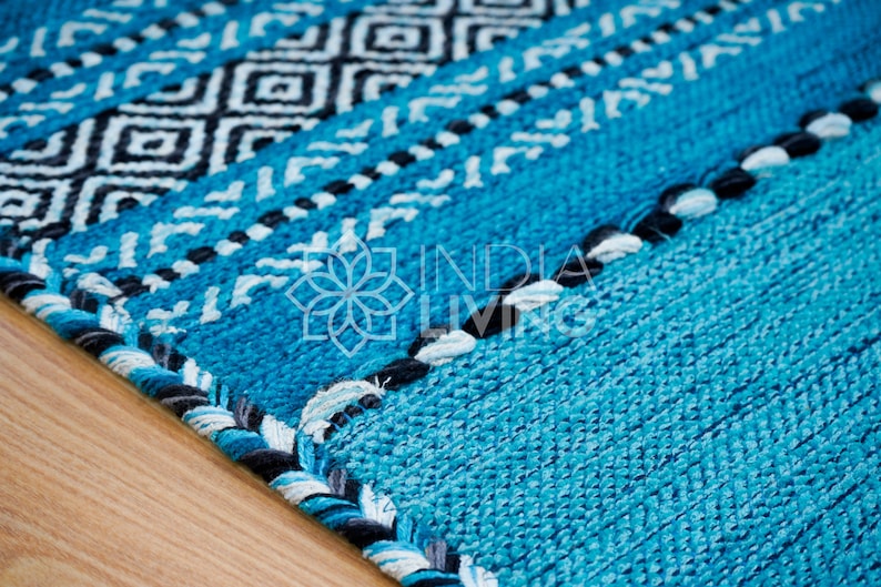 May include: A woven rug featuring a vibrant turquoise blue base with alternating bands of black and white geometric diamond patterns and braided trim. The center of the rug displays the white logo text INDIA LIVING with a floral icon above it.