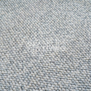 May include: A close-up of a gray and white round rug made of small, round, felt balls. The rug has a subtle pattern of alternating colors.