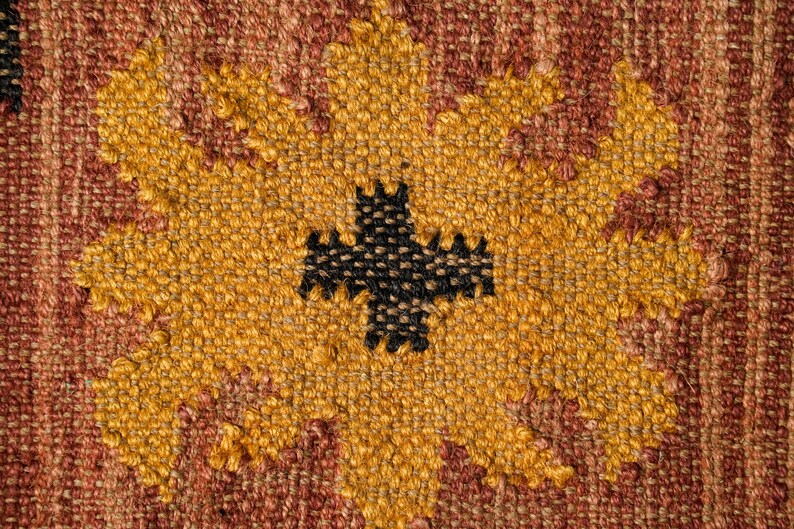May include: Close-up of a woven fabric with a yellow and brown pattern. The pattern features a large, stylized flower with a black cross in the center.