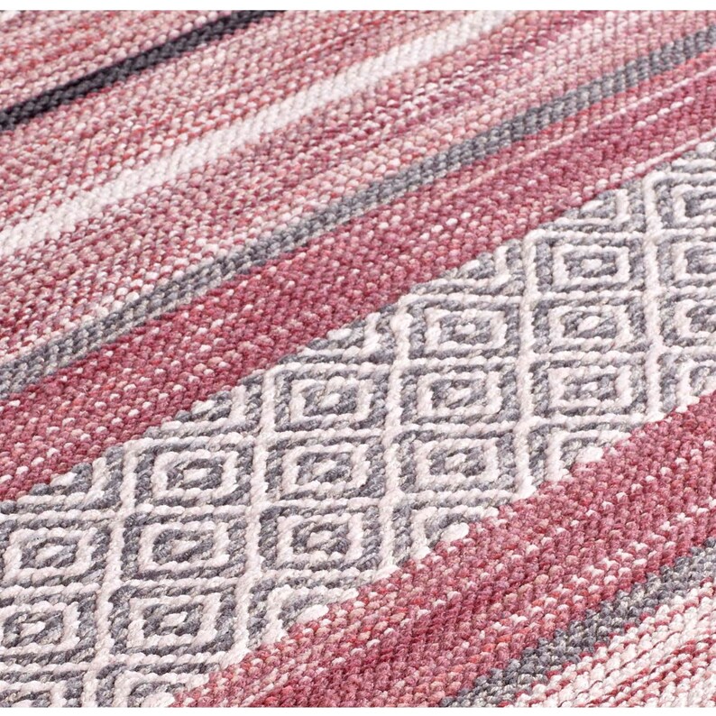 May include: Close-up of a woven rug with a geometric pattern. The rug features a combination of colors, including shades of red, gray, and white. Diagonal stripes and diamond shapes create a textured surface, suitable for home decor.