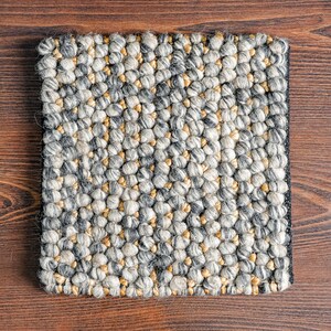 Puede incluir: Una alfombra cuadrada con una superficie densa y texturizada compuesta por numerosas peque&ntilde;as bolas de lana afieltrada redondeadas en tonos gris carb&oacute;n, gris claro, crema y beige, dispuestas en un patr&oacute;n repetitivo con un borde oscuro.