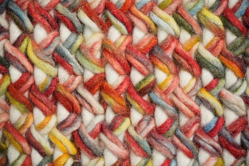 May include: A close-up view of a thick, braided textile featuring a repeating pattern of chunky, off-white yarn strands interwoven with variegated yarn in shades of red, orange, yellow, green, and blue, creating a dense, textured surface.