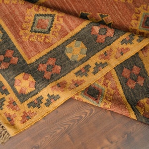 Puede incluir: Una alfombra tejida con un patr&oacute;n geom&eacute;trico en tonos marrones, naranjas y amarillos. La alfombra tiene un borde de flecos y est&aacute; doblada por la mitad.