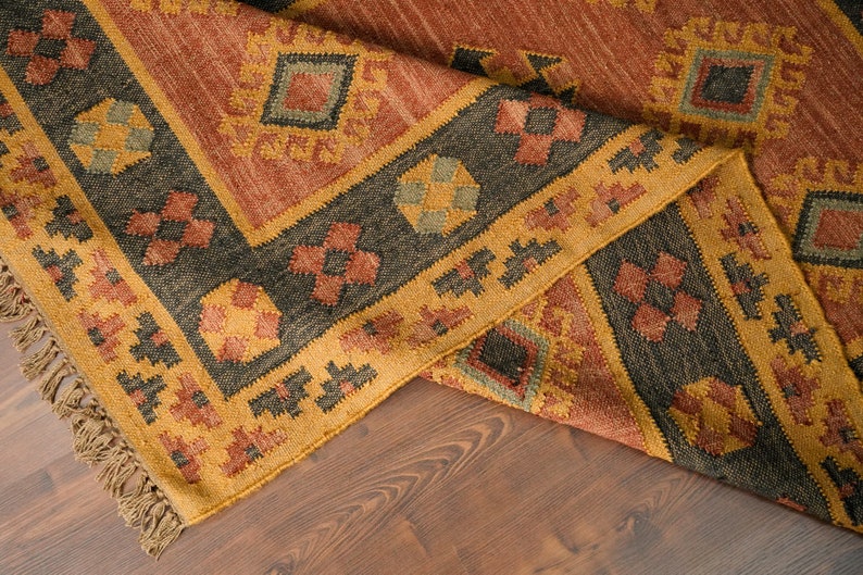 Puede incluir: Una alfombra tejida con un patr&oacute;n geom&eacute;trico en tonos marrones, naranjas y amarillos. La alfombra tiene un borde de flecos y est&aacute; doblada por la mitad.