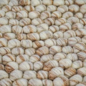 K&ouml;nnte beinhalten: Nahaufnahme eines runden, beige-braunen Wollteppichs aus kleinen, runden Kugeln. Der Teppich hat eine strukturierte, flauschige Oberfl&auml;che.