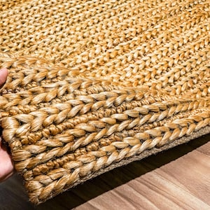 May include: Close-up of a natural fiber rug with a braided border. The rug is a warm, light brown color, with a textured surface. The braided border adds a decorative element to the rug. The rug is made of a durable material.