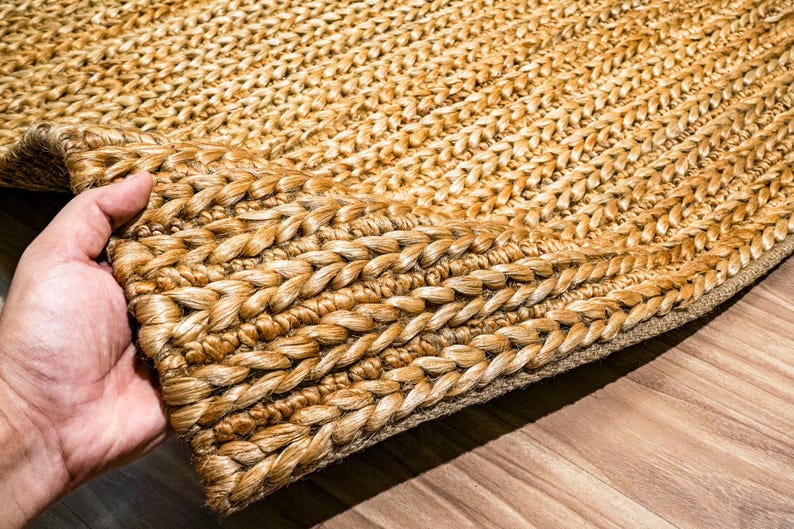 May include: Close-up of a natural fiber rug with a braided border. The rug is a warm, light brown color, with a textured surface. The braided border adds a decorative element to the rug. The rug is made of a durable material.