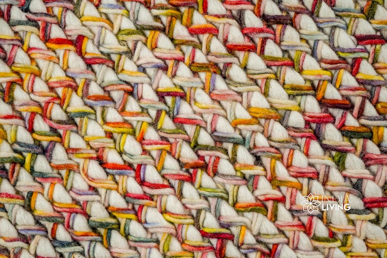 May include: A thick, hand-woven textile featuring a dense, braided pattern of cream-colored yarn interlaced with multicolored strands in shades of red, yellow, green, and blue. The surface displays a repeating diagonal weave with the text INDIA LIVING.