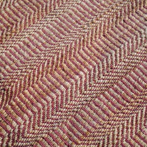 May include: Close-up of a woven rug with a pink and beige chevron pattern.