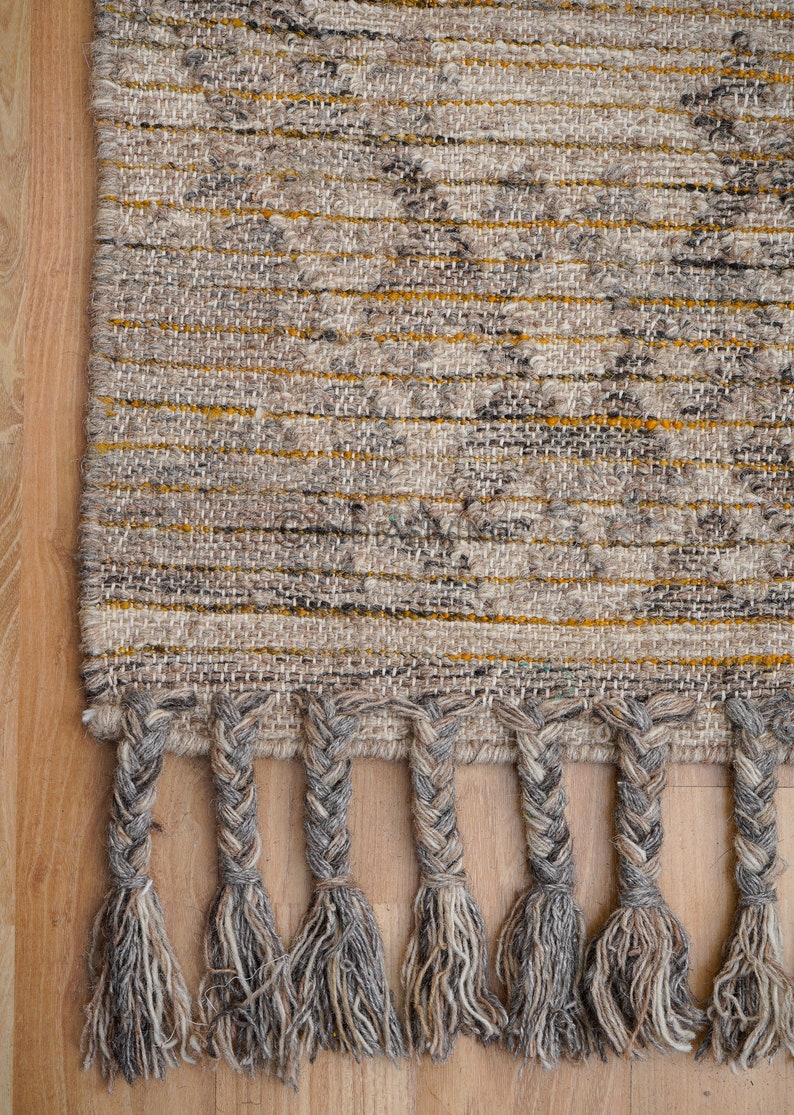 May include: A woven rug with a gray and yellow geometric pattern and braided fringe. The rug is made of natural fibers and has a rustic, hand-crafted look.