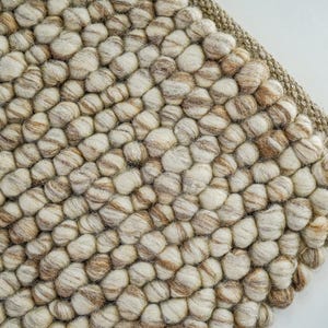 K&ouml;nnte beinhalten: Nahaufnahme eines beige-braunen Wollteppichs mit einer strukturierten, unebenen Oberfl&auml;che. Der Teppich besteht aus kleinen, runden Wollkugeln, die miteinander verwoben sind.