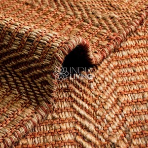 May include: A woven rug featuring a textured herringbone pattern in alternating shades of natural tan and burnt orange. The surface displays a thick, braided fiber construction with a visible fold, and the text INDIA LIVING is printed in white.