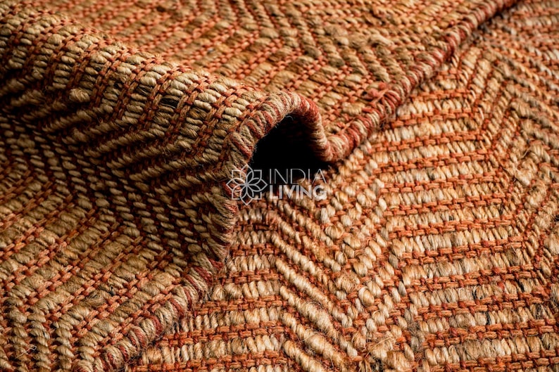 May include: A woven rug featuring a textured herringbone pattern in alternating shades of natural tan and burnt orange. The surface displays a thick, braided fiber construction with a visible fold, and the text INDIA LIVING is printed in white.