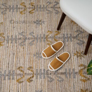 May include: A beige and gray patterned rug with a subtle geometric design. Two brown slippers with white trim are on the rug. The rug is in a room with a white chair and a green plant.
