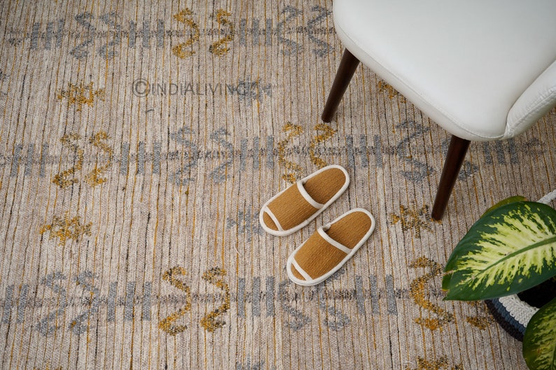 May include: A beige and gray patterned rug with a subtle geometric design. Two brown slippers with white trim are on the rug. The rug is in a room with a white chair and a green plant.