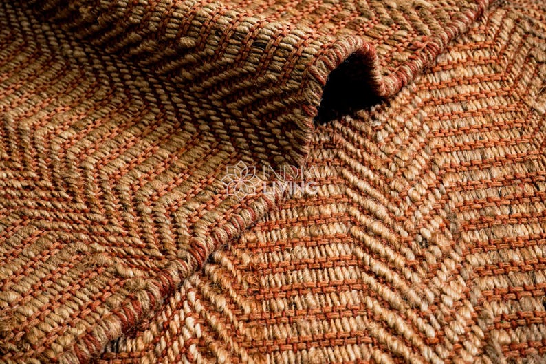 May include: A woven rug featuring a textured herringbone pattern in alternating shades of natural tan and burnt orange. The material appears to be a thick, fibrous natural fiber with a visible weave, showing a folded corner revealing the underside.