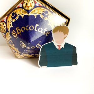 Pode incluir: Uma caixa azul escura com detalhes dourados e a palavra "Chocolate" em dourado. Um adesivo de um personagem com um su&eacute;ter verde e gravata vermelha est&aacute; na frente da caixa. A caixa tem formato de diamante.