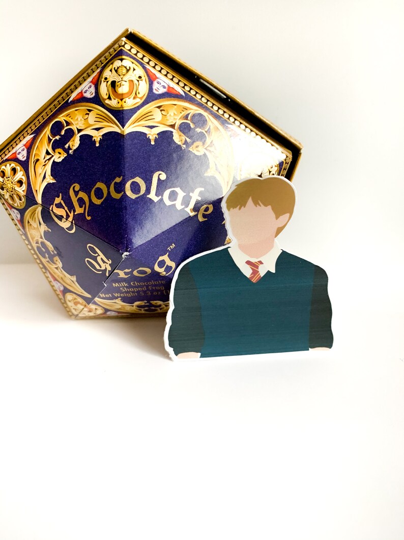 Pode incluir: Uma caixa azul escura com detalhes dourados e a palavra "Chocolate" em dourado. Um adesivo de um personagem com um su&eacute;ter verde e gravata vermelha est&aacute; na frente da caixa. A caixa tem formato de diamante.