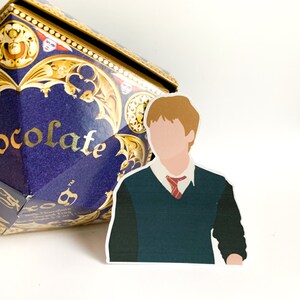 Pode incluir: Um adesivo de um personagem com cabelo ruivo, usando um su&eacute;ter verde, camisa branca e gravata vermelha. Uma caixa azul e dourada com a palavra "chocolate" est&aacute; ao fundo.