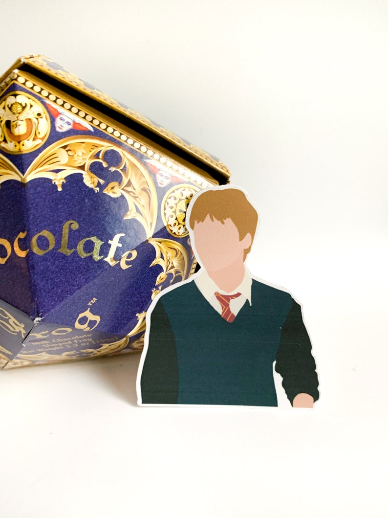 Pode incluir: Um adesivo de um personagem com cabelo ruivo, usando um su&eacute;ter verde, camisa branca e gravata vermelha. Uma caixa azul e dourada com a palavra "chocolate" est&aacute; ao fundo.