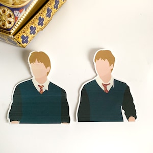Pode incluir: Dois adesivos com um personagem em uniforme escolar com um su&eacute;ter verde, camisa branca e gravata vermelha. Os adesivos t&ecirc;m um design minimalista com cabelo castanho claro e sem tra&ccedil;os faciais. Uma caixa decorativa est&aacute; no fundo.