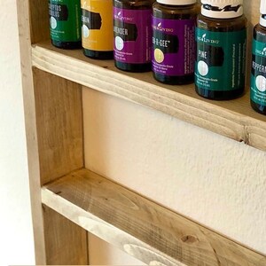 May include: A wooden shelf with a driftwood stain finish, holding rows of essential oil bottles with white lids. The bottles are labeled with the brand "Young Living" and the names of the oils, including "Eucalyptus Globulus", "Lemon", "Lavender", "En-R-Gee", "Pine", and "Peppermint".