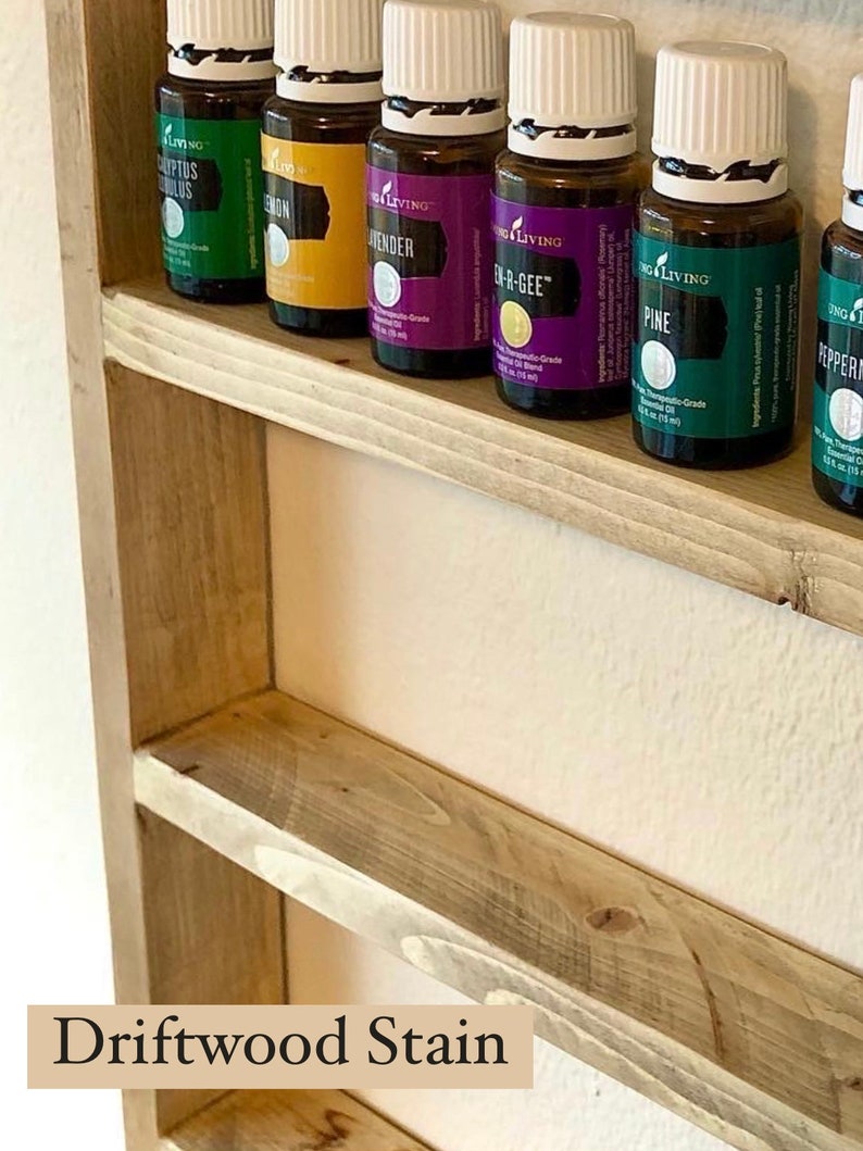 May include: A wooden shelf with a driftwood stain finish, holding rows of essential oil bottles with white lids. The bottles are labeled with the brand "Young Living" and the names of the oils, including "Eucalyptus Globulus", "Lemon", "Lavender", "En-R-Gee", "Pine", and "Peppermint".