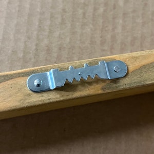 May include: A silver metal hanging hook with a sawtooth design, attached to a wooden frame.