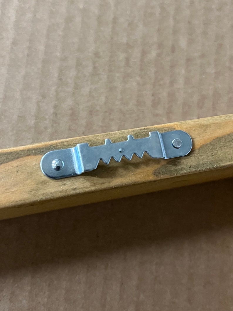 May include: A silver metal hanging hook with a sawtooth design, attached to a wooden frame.