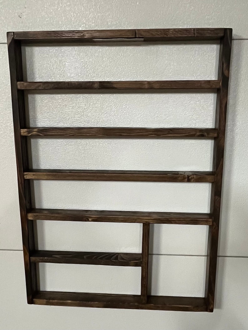 May include: A dark brown wooden wall shelf with five shelves and a small cubby on the bottom right. The shelf is mounted on a white wall.