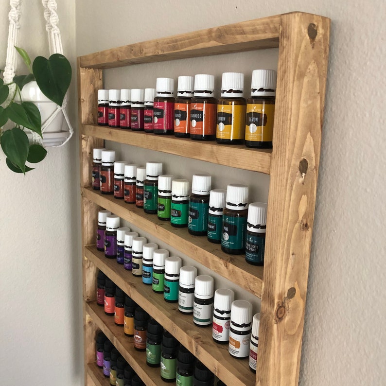 May include: A wooden wall shelf with multiple shelves, each filled with small glass bottles with white lids. The bottles are arranged in rows and are filled with colorful liquids. The shelf is made of natural wood and has a rustic look.